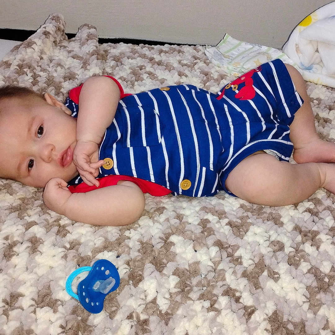 Asher is registered to the contest to win money with this photo: baby, blanket, blue, child, clothing, comfort, cute, floor, indoor, infant, knit, lying_down, nursery, pacifier, plush_toy, red, relaxed, romper, soft, white