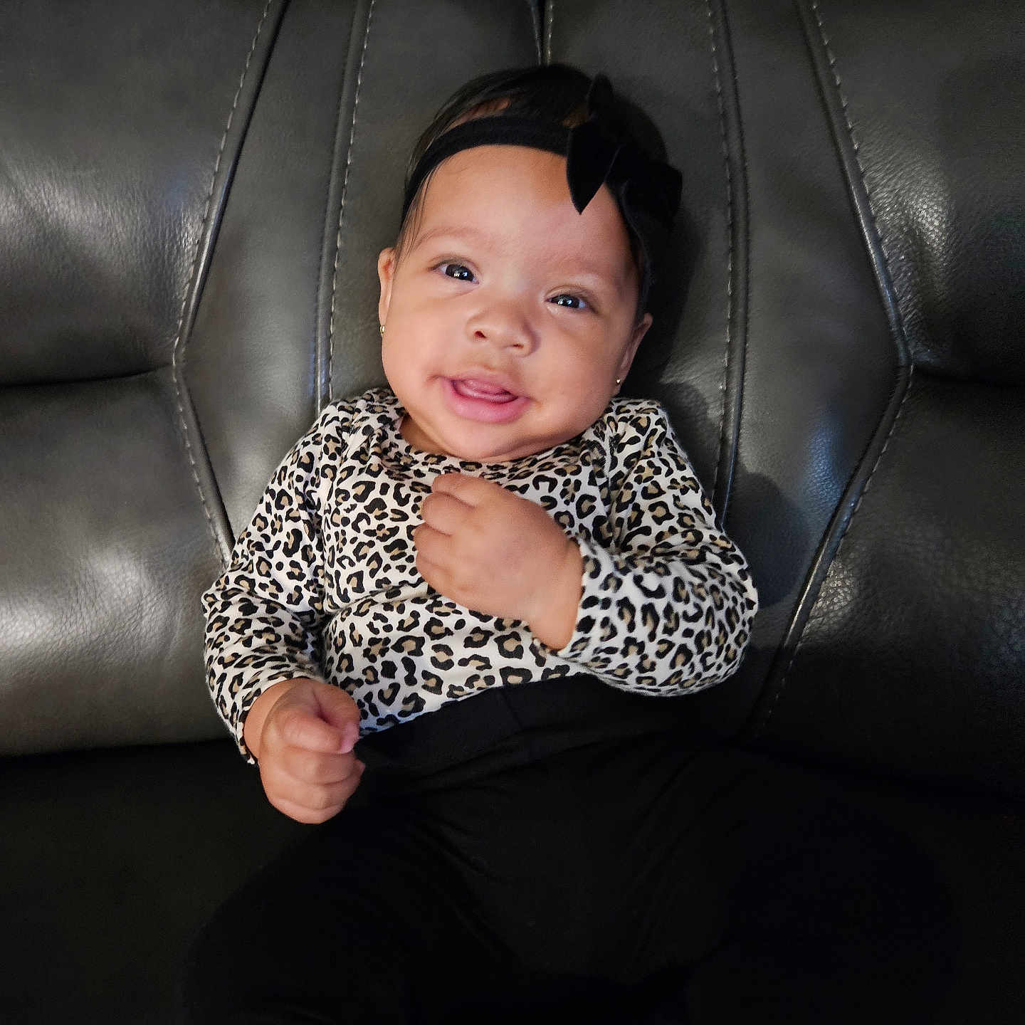 Kaveri is registered to the contest to win money with this photo: baby, child, infant, smiling, sitting, portrait, leopard_print, outfit, black_bow, headband, leather_couch, furniture, indoors, close_up, face, hands, dark_clothing, happy, adorable, photography