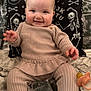 baby, child, smiling, ribbed_clothing, beige_outfit, blanket, pacifier, pillow, skeleton_design, celestial_symbols, indoor, portrait, happy, seated, cute, infant, face, hand, cozy, soft_fabric