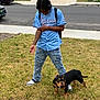 person, dog, grass, street, sidewalk, jeans, tattoo, headphones, backpack, car, house, lawn, black_dog, blue_jersey, urban, outdoor, casual, short_hair, standing, alert