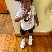 Gemario is registered to the contest to win money with this photo: child, boy, cap, nike, white_shirt, black_shorts, chains, wooden_floor, pointing, sneakers, white_sneakers, expression, indoor, serious, flooring, standing, person, footwear, casual, wall