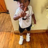 child, boy, cap, nike, white_shirt, black_shorts, chains, wooden_floor, pointing, sneakers, white_sneakers, expression, indoor, serious, flooring, standing, person, footwear, casual, wall