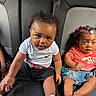 child, toddler, car_seat, baby, clothing, hair_clips, denim_skirt, white_shirt, black_shorts, socks, seatbelt, curly_hair, indoor, portrait, two_children, cute, looking_at_camera, infant, casual, car_interior