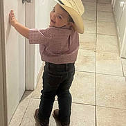 Colton is registered to the contest to win money with this photo: child, boy, hat, cowboy_hat, smile, shirt, jeans, shoes, indoor, hallway, tile_floor, door, wall, standing, looking_back, playful, young, person, casual_clothing, cute