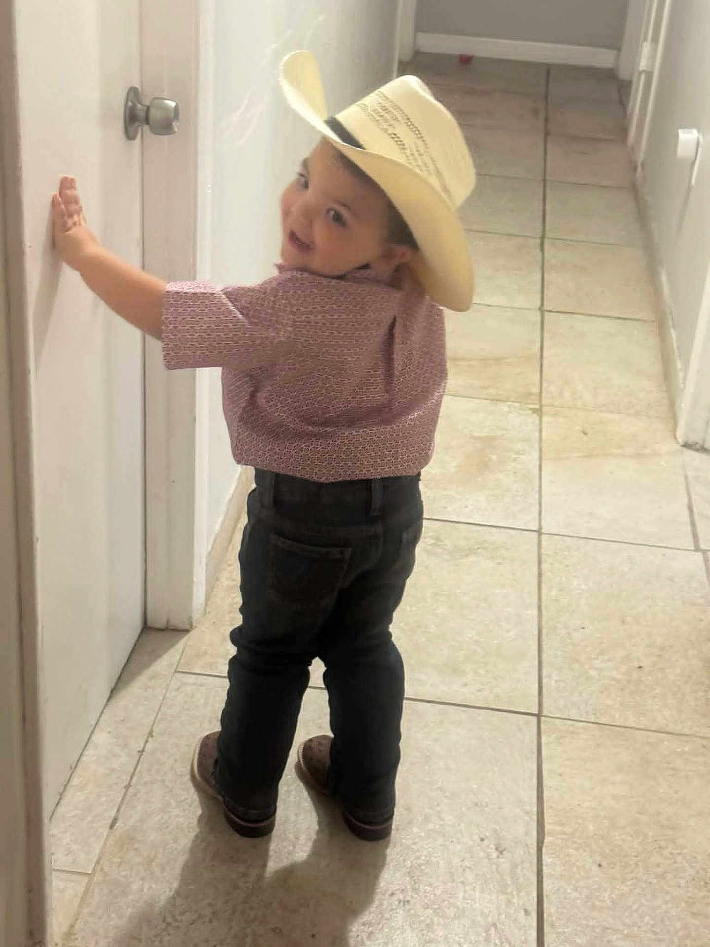 Colton is registered to the contest to win money with this photo: child, boy, hat, cowboy_hat, smile, shirt, jeans, shoes, indoor, hallway, tile_floor, door, wall, standing, looking_back, playful, young, person, casual_clothing, cute