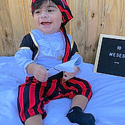 Neizan Ramírez joined the competition — help win amazing prizes! baby, child, pirate_costume, smiling, sitting, white_blanket, wooden_fence, red_and_black, hat, pants, black_socks, paper, necklace, outdoor, happy, portrait, cute, young_child, costume, playful