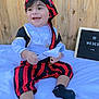 baby, child, pirate_costume, smiling, sitting, white_blanket, wooden_fence, red_and_black, hat, pants, black_socks, paper, necklace, outdoor, happy, portrait, cute, young_child, costume, playful