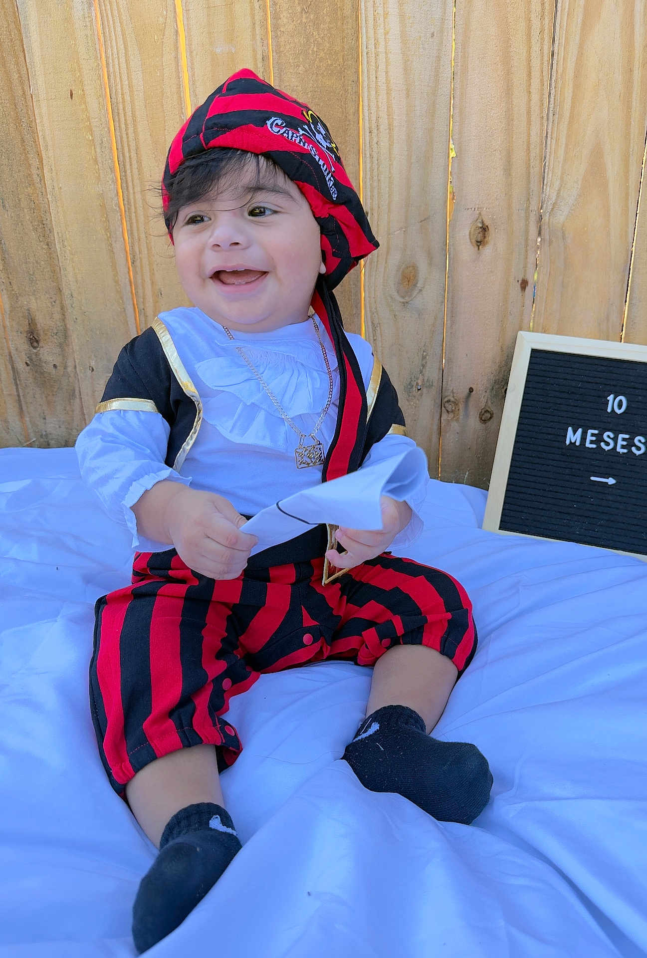 Neizan Ramírez joined the competition — help win amazing prizes! baby, child, pirate_costume, smiling, sitting, white_blanket, wooden_fence, red_and_black, hat, pants, black_socks, paper, necklace, outdoor, happy, portrait, cute, young_child, costume, playful