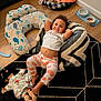 child, toddler, smiling, indoor, bean_bag, doll, pet_bed, dog, floor, rug, pillow, clothing, toy, wood_floor, pet_bowl, blanket, happy, resting, home, casual