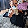 toddler, child, milk_bottle, box, curly_hair, carpet, bed, pillow, indoor, window, smiling, sitting, furniture, blanket, floor, curtain, person, happy, room, cozy