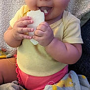 Aria is registered to the contest to win money with this photo: baby, child, smiling, eating, bread, blanket, yellow_clothing, pink_clothing, happy, indoor, cute, infant, person, face, hands, seated, cozy, soft_texture, closeup, portrait