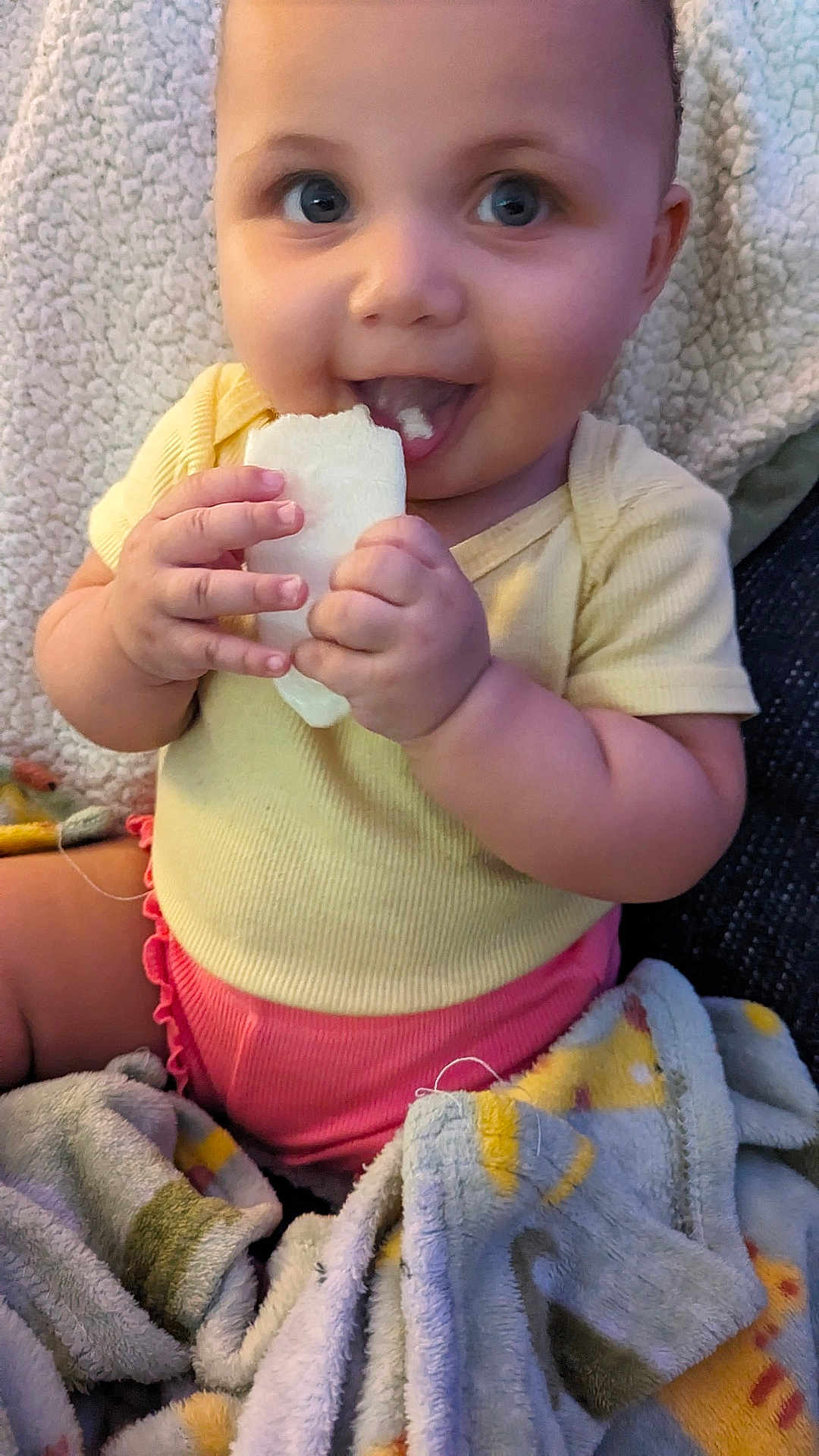 Aria is registered to the contest to win money with this photo: baby, child, smiling, eating, bread, blanket, yellow_clothing, pink_clothing, happy, indoor, cute, infant, person, face, hands, seated, cozy, soft_texture, closeup, portrait
