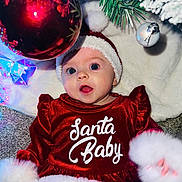 Kiera is registered to the contest to win money with this photo: baby, christmas, holiday, red_dress, santa_hat, ornament, christmas_tree, decorations, festive, infant, face, blue_eyes, carpet, white_fur_trim, celebration, smiling, cute, seasonal, indoor, portrait