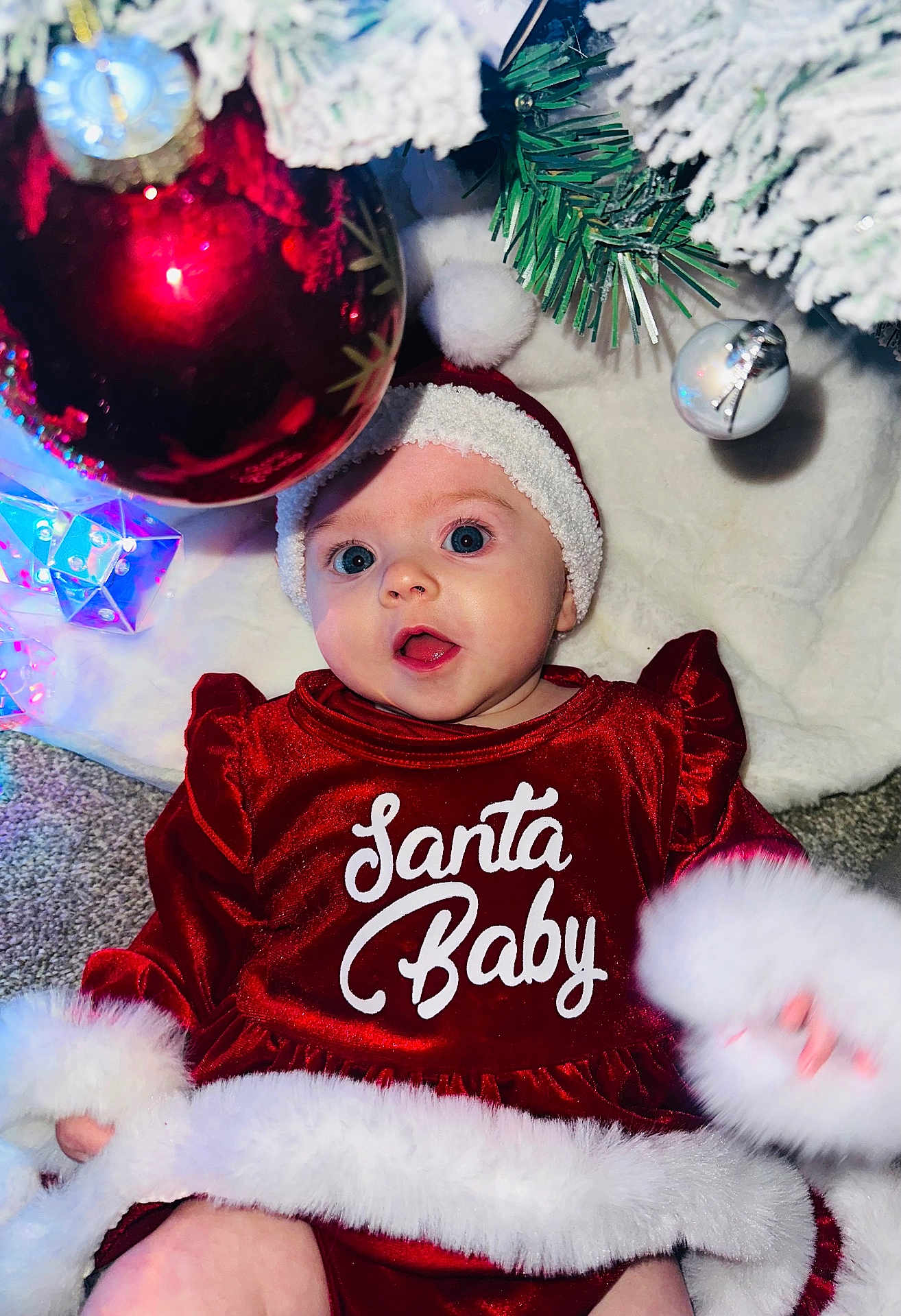 Kiera is registered to the contest to win money with this photo: baby, christmas, holiday, red_dress, santa_hat, ornament, christmas_tree, decorations, festive, infant, face, blue_eyes, carpet, white_fur_trim, celebration, smiling, cute, seasonal, indoor, portrait