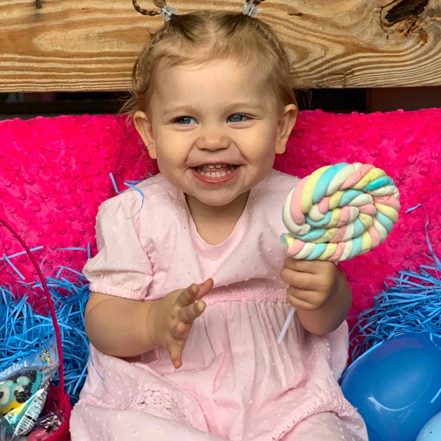 Haylie is registered to the contest to win money with this photo: basket, blue_shredded_paper, braided_hair, candy, celebration, child, cute, easter_eggs, face, food, happy, holiday, indoors, lollipop, pink_blanket, pink_dress, playful, sitting, smiling, toddler