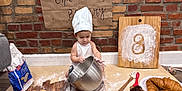 Enki is registered to the contest to win money with this photo: baby, child, chef_hat, flour, baking, bread, croissants, muffins, mixing_bowl, whisk, wooden_board, brick_wall, table, apron, kitchen_utensils, pastry, homemade, food, cooking, cute