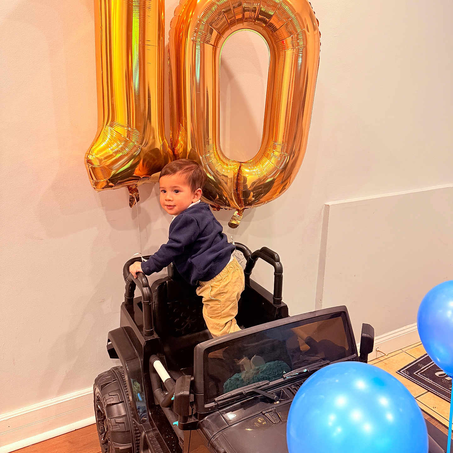 Enki joined the competition — help win amazing prizes! balloons, blue_balloons, celebration, child, clothing, floor, gold_balloons, happy, indoor, number_10, pants, party, person, smiling, sweater, toddler, toy_car, wall, window, wooden_floor