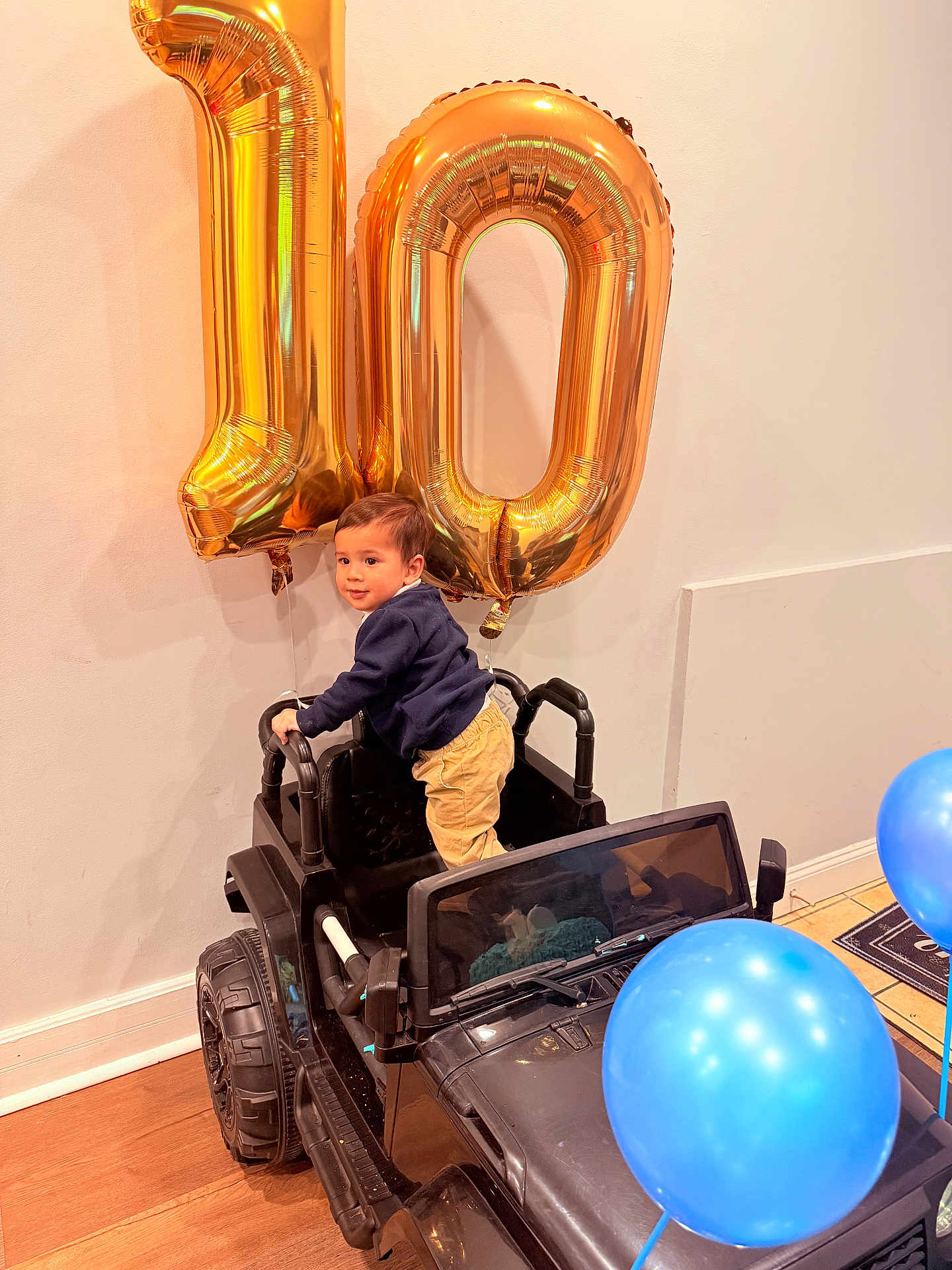 Enki joined the competition — help win amazing prizes! toddler, child, toy_car, balloons, gold_balloons, number_10, indoor, wooden_floor, blue_balloons, celebration, party, smiling, person, clothing, pants, sweater, floor, wall, window, happy