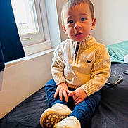 Enki joined the competition — help win amazing prizes! child, toddler, sitting, sweater, pants, shoes, bed, window, curtain, indoor, natural_light, expression, face, hands, footwear, cozy, clothing, person, young_child, curious