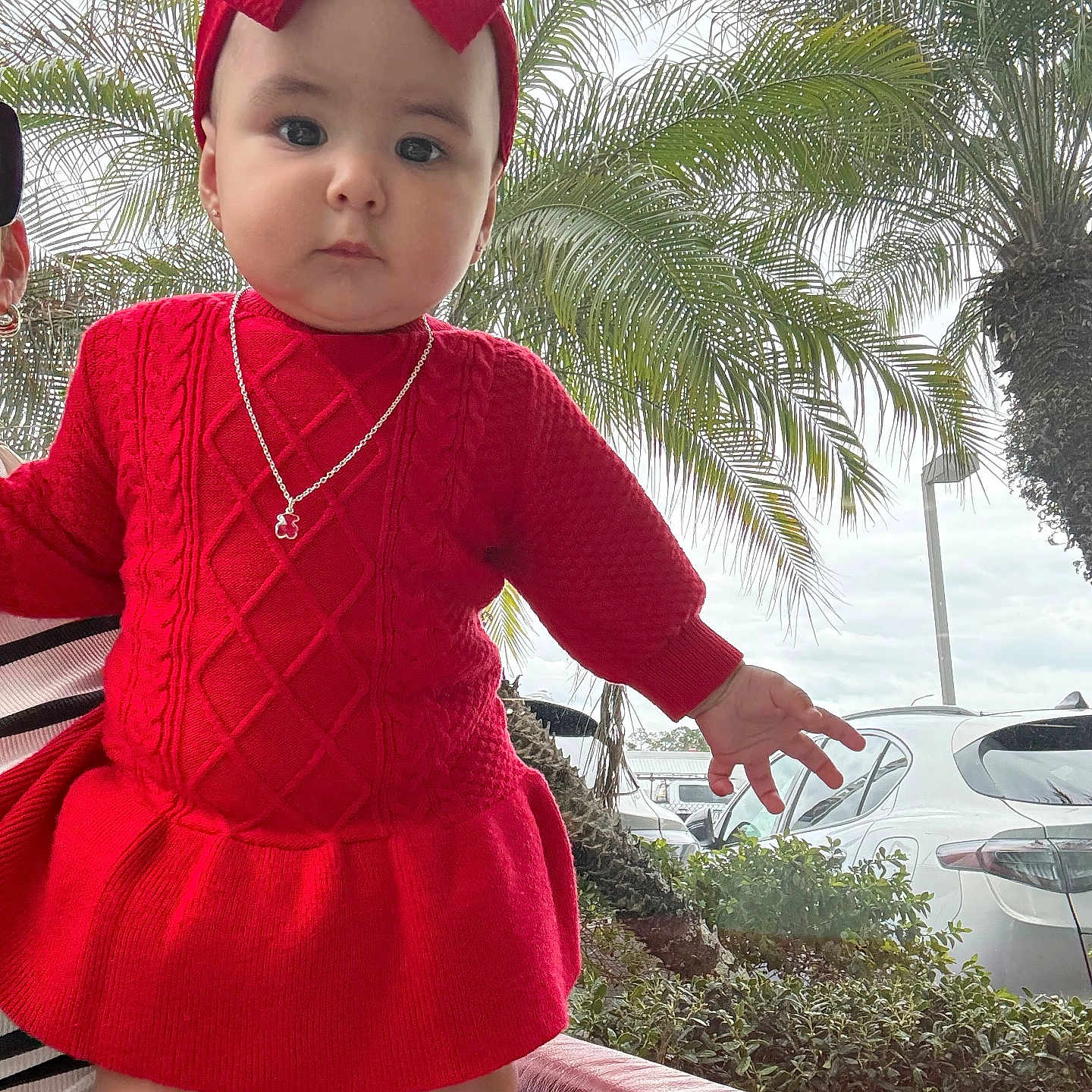 Daireliz is registered to the contest to win money with this photo: baby, car, child, cute, daytime, earrings, fashion, hand, infant, large_bow, necklace, outdoor, palm_trees, parking_lot, plants, portrait, red_chair, red_dress, serious_expression, sitting