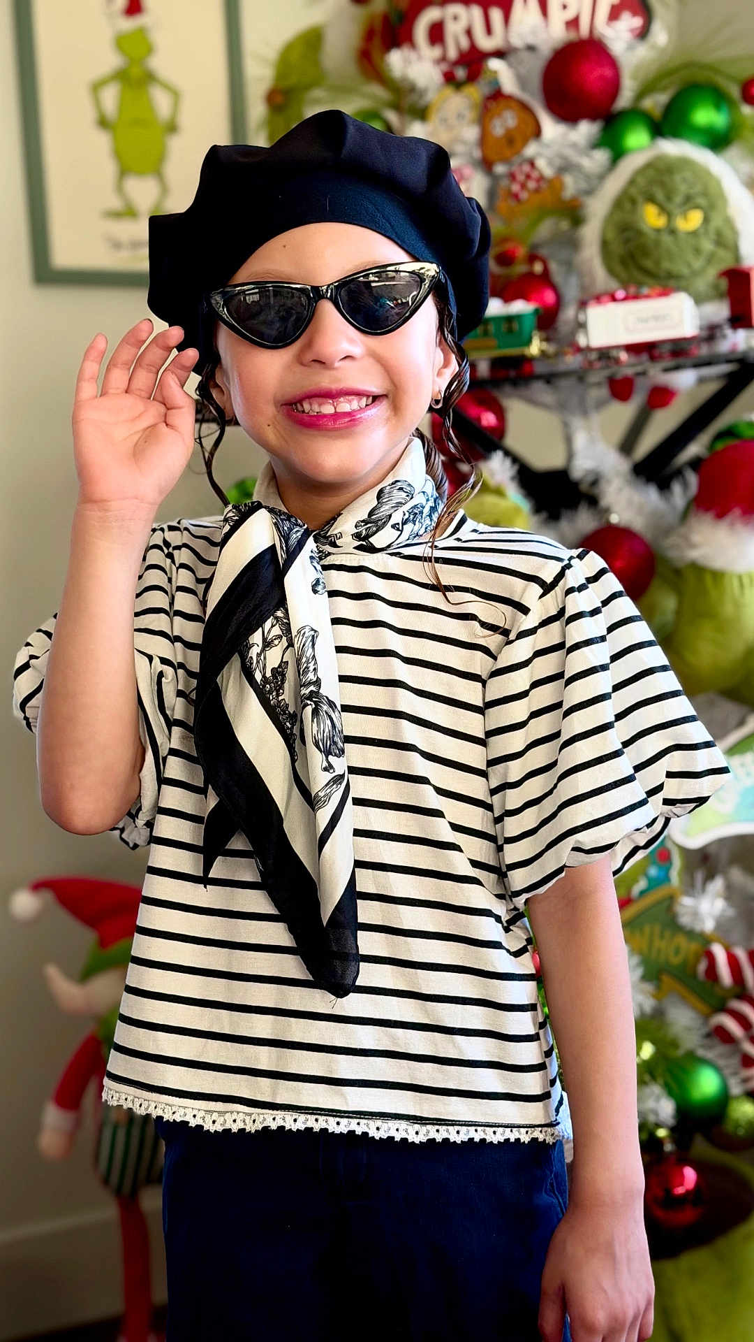 Samantha is registered to the contest to win money with this photo: child, smiling, beret, sunglasses, striped_shirt, scarf, holiday_decorations, grinch, christmas, festive, indoors, portrait, hand_wave, happy, fashion, accessories, toy, decor, person, cute