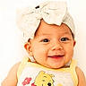 baby, child, smiling, headband, bow, yellow_clothing, cartoon_character, face, portrait, happy, cute, infant, closeup, light_background, cheerful, young_child, apparel, person, indoors, headwear