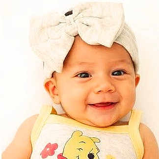 Emily is registered to the contest to win money with this photo: baby, child, smiling, headband, bow, yellow_clothing, cartoon_character, face, portrait, happy, cute, infant, closeup, light_background, cheerful, young_child, apparel, person, indoors, headwear
