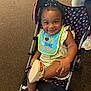 Maliya joined the competition — help win amazing prizes! toddler, child, smile, stroller, bib, footwear, happy, indoor, carpet, person, clothing, sandals, baby, cute, seated, playful, hair, face, expression, accessory