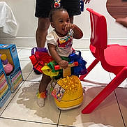 Maliya joined the competition — help win amazing prizes! child, toddler, toy, ride_on_toy, yellow, rainbow_tutu, colorful, indoor, red_chair, person, floor_tiles, smiling, playful, adult_legs, gift_bow, hand, happy, clothing, hair_accessory, sock