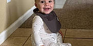 Caden is registered to the contest to win money with this photo: baby, child, sitting, smiling, pajamas, bib, floor, tile_floor, carpet, indoor, home, door, wall, happy, portrait, person, cute, young, infant, clothing