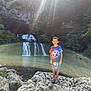 child, boy, waterfall, rocks, rainbow, nature, outdoor, forest, sunlight, tshirt, shorts, sandals, landscape, scenic, water, smile, standing, daylight, clothing, recreation
