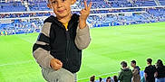 Thiago participe au concours pour gagner de l'argent avec cette photo : child, boy, stadium, soccer_field, seats, spectators, sports_event, casual_clothing, peace_sign, smiling, outdoor, daytime, blue_seats, jeans, sneakers, crowd, photography, person, active, fun