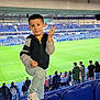 child, boy, stadium, soccer_field, seats, spectators, sports_event, casual_clothing, peace_sign, smiling, outdoor, daytime, blue_seats, jeans, sneakers, crowd, photography, person, active, fun