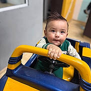 Liam is registered to the contest to win money with this photo: toddler, child, toy_car, indoor, yellow, blue, handlebar, steering_wheel, curious, short_hair, green_clothing, playing, young_child, portrait, focus, hand, expression, floor, background, room