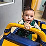 toddler, child, toy_car, indoor, yellow, blue, handlebar, steering_wheel, curious, short_hair, green_clothing, playing, young_child, portrait, focus, hand, expression, floor, background, room