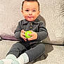 Liam is registered to the contest to win money with this photo: baby, child, smiling, sitting, toy, green_toy, gray_couch, gray_clothing, socks, cute, indoors, portrait, happy, hands, face, hair, infant, cozy, casual, playful