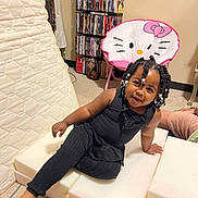 Myayla is registered to the contest to win money with this photo: child, toddler, girl, portrait, smiling, sitting, indoor, playroom, black_outfit, braided_hair, hair_beads, hello_kitty, pink_chair, ottoman, bookshelf, movies, mattress, carpet, barefoot, home