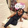 child, toddler, girl, portrait, smiling, sitting, indoor, playroom, black_outfit, braided_hair, hair_beads, hello_kitty, pink_chair, ottoman, bookshelf, movies, mattress, carpet, barefoot, home