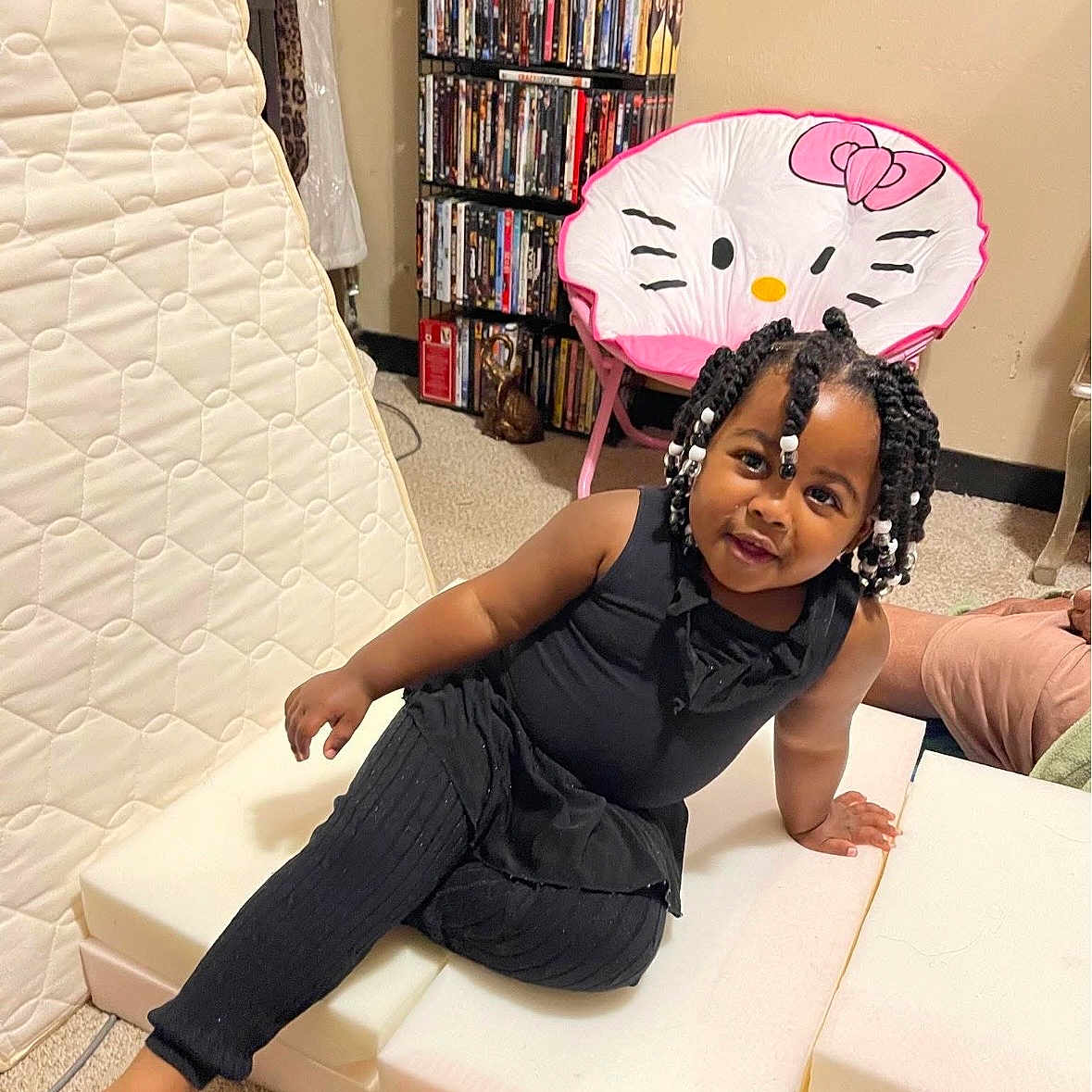 Myayla is registered to the contest to win money with this photo: child, toddler, girl, portrait, smiling, sitting, indoor, playroom, black_outfit, braided_hair, hair_beads, hello_kitty, pink_chair, ottoman, bookshelf, movies, mattress, carpet, barefoot, home