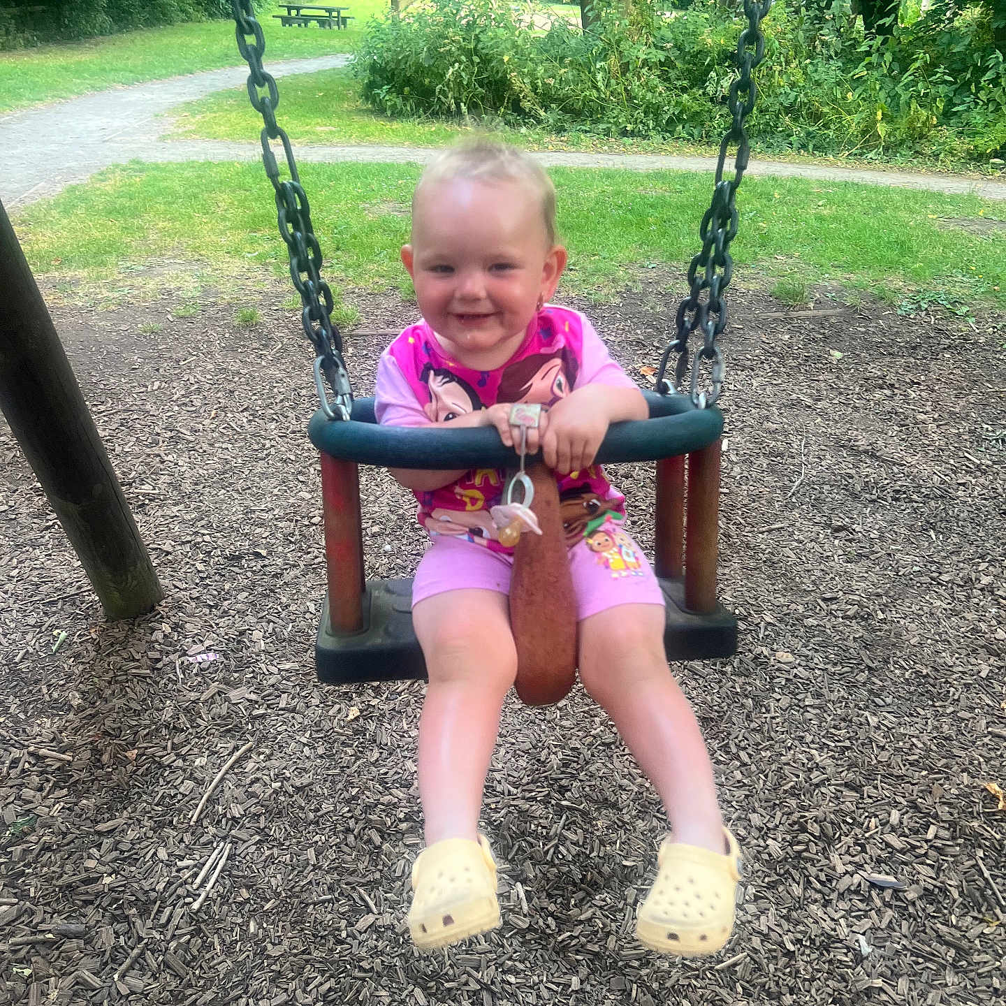 Olivia Mae joined the competition — help win amazing prizes! baby, child, daylight, fun, grass, greenery, happy, outdoor, pacifier, park, person, pink_pajamas, play, playground, smile, swing, toddler, trees, woodchips, yellow_crocs