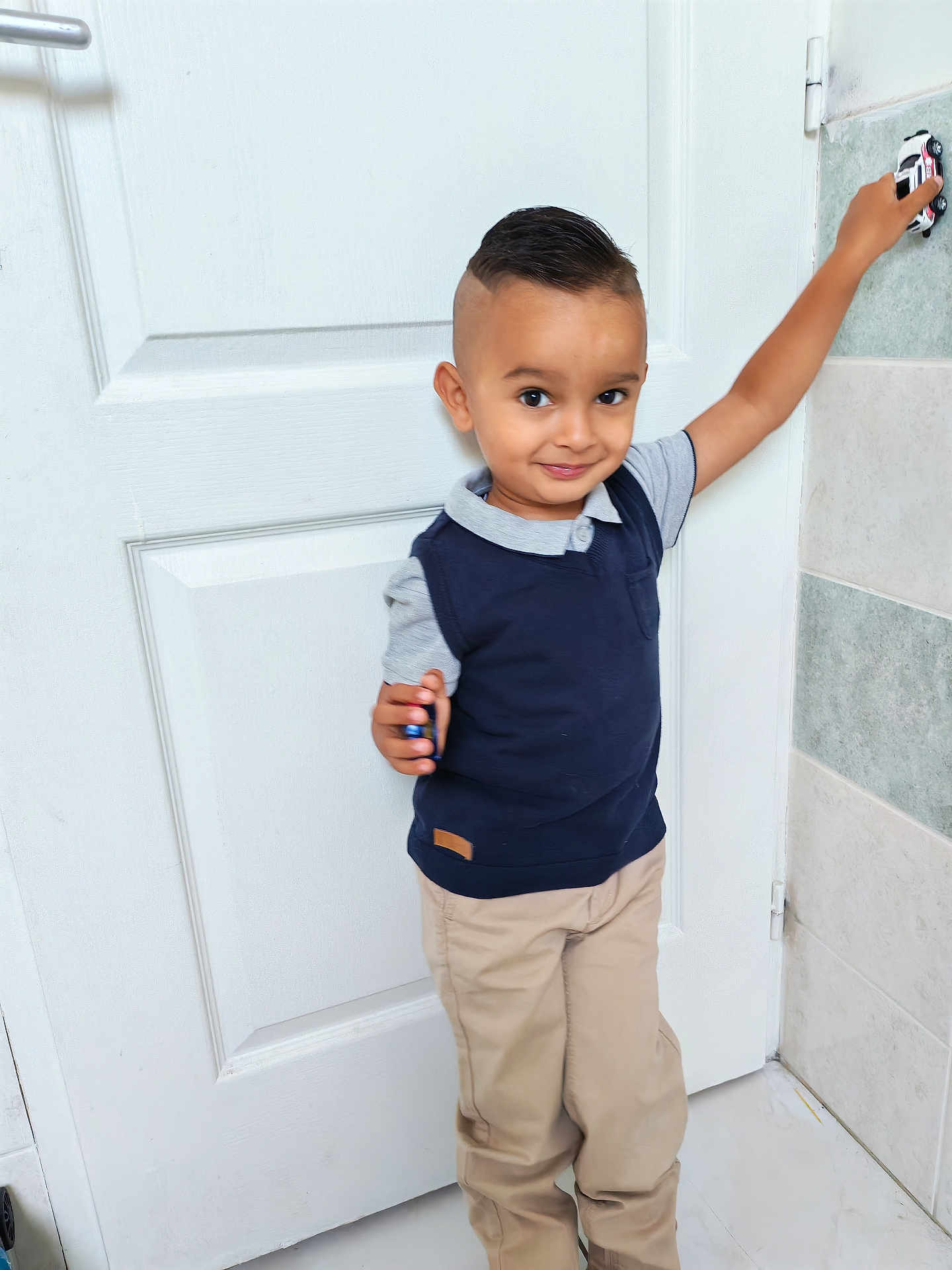 Melkior participe au concours pour gagner de l'argent avec cette photo : child, boy, toy_car, door, indoor, smile, short_hair, casual_clothing, pants, shirt, vest, playful, standing, hand, wall, floor, portrait, cute, person, young