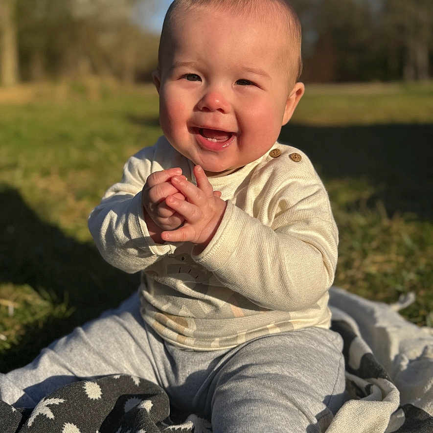 James is registered to the contest to win money with this photo: baby, blanket, casual_clothing, child, clapping, cute, daylight, face, grass, happy, head, infant, nature, outdoor, playful, portrait, sitting, smiling, sunlight, young