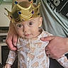 baby, child, crown, onesie, sloth_pattern, adult_hand, indoor, curious, wide_eyes, portrait, cute, jewels, headwear, holding, person, clothing, face, expression, young_child, family