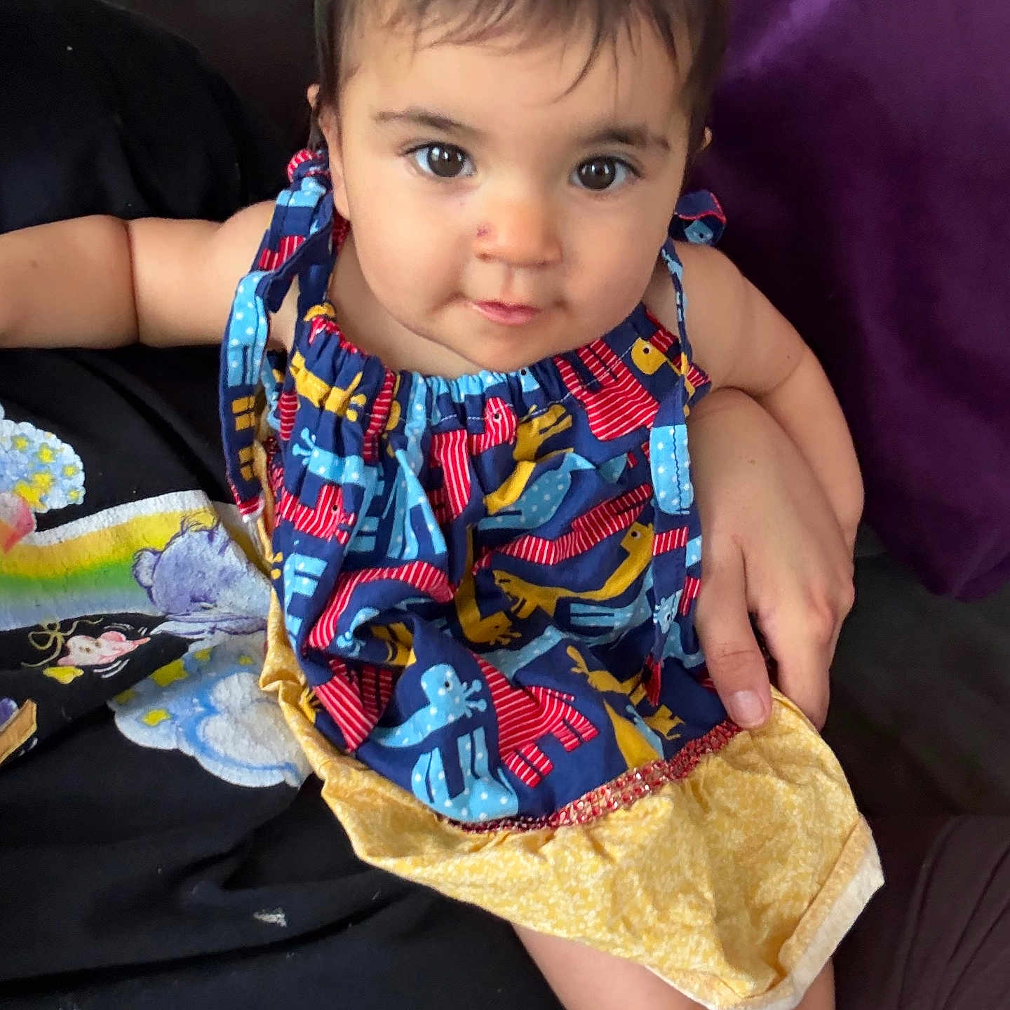 Melody is registered to the contest to win money with this photo: baby, toddler, child, person, face, eyes, dress, colorful_clothing, sitting, arm, hand, foot, indoor, couch, adult, holding, purple_background, black_clothing, cute, portrait