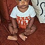 baby, child, sitting, couch, orange, white_shirt, curious, indoors, face, skin, hand, foot, pillow, textile, seated, infant, young_child, portrait, closeup, furniture