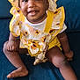 baby, smiling, child, yellow_clothing, bonnet, sunflower_pattern, crocheted_toy, blue_couch, sitting, happy, cute, infant, indoors, portrait, face, person, young_child, playful, cheerful, clothing
