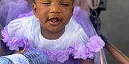 Ily a rejoint le concours — aidez-le/la à gagner de superbes lots ! baby, child, stroller, water_bottle, purple_dress, white_shirt, tutu, toy, blanket, hand, face, expression, outdoor, seat, infant, clothing, cute, reaching, playful, fabric
