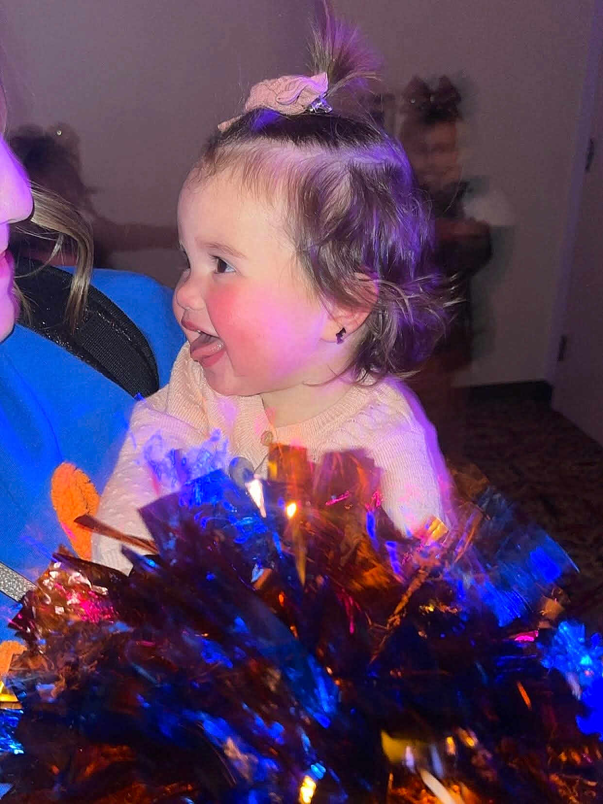 Brae joined the competition — help win amazing prizes! toddler, child, person, face, hair, pony_tail, smile, tongue, cheeks, pom_poms, colorful, blue_light, orange_light, indoor, blurred_background, happy, joyful, holding, clothing, playful