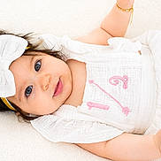 Liliana joined the competition — help win amazing prizes! baby, infant, girl, white_clothing, headband, bow, blue_eyes, bracelet, lying_down, soft_surface, portrait, cute, smiling, child, young, pink_embroidery, skin, fashion, adorable, closeup