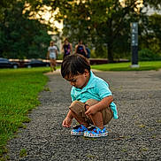 Malachi is registered to the contest to win money with this photo: child, toddler, boy, squatting, blue_crocs, turquoise_shirt, shorts, outdoor, park, path, grass, trees, people, background, daylight, curious, playing, casual_clothing, nature, shoes